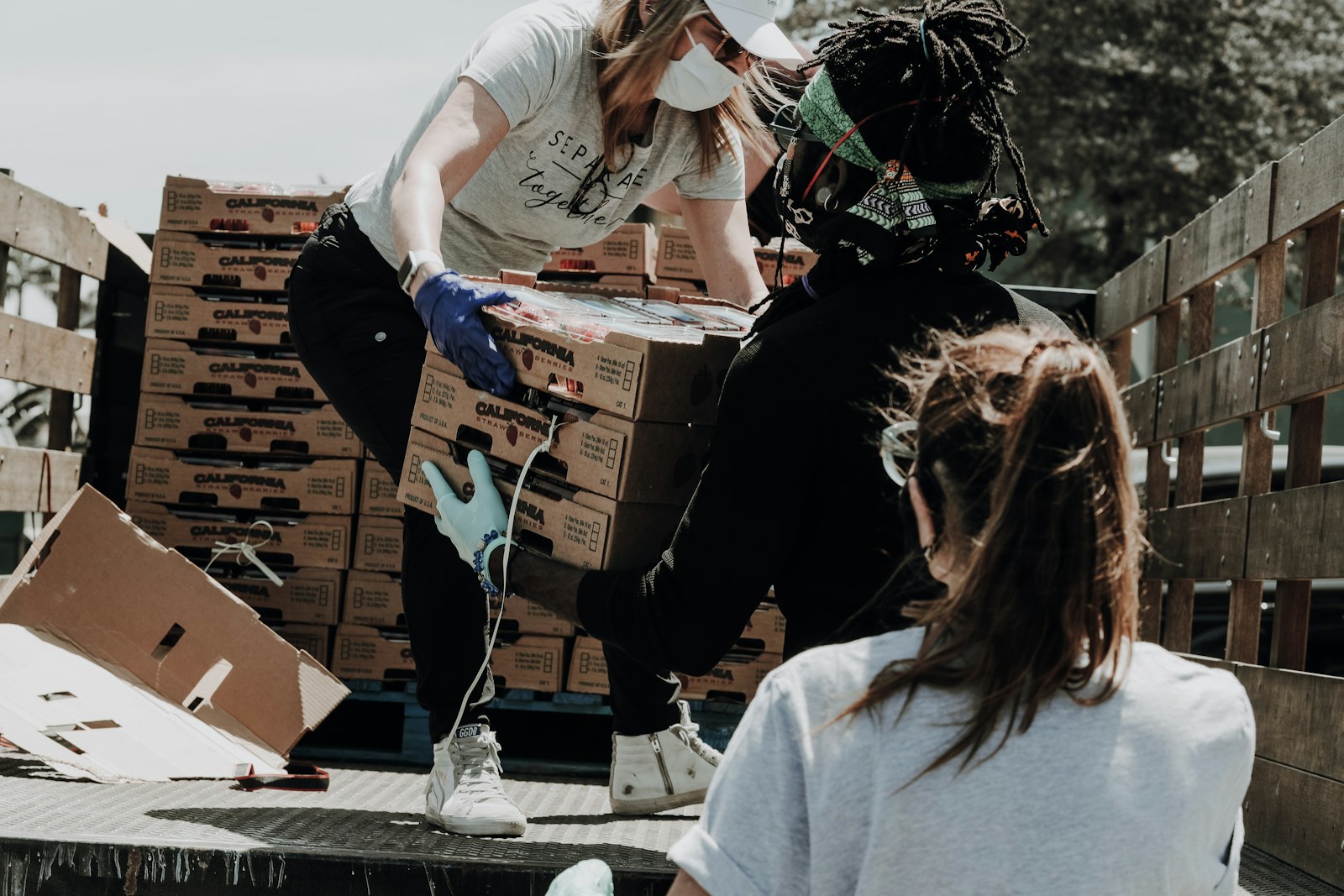 Three people wearing masks loading cardboard boxes of strawberries onto a truck bed