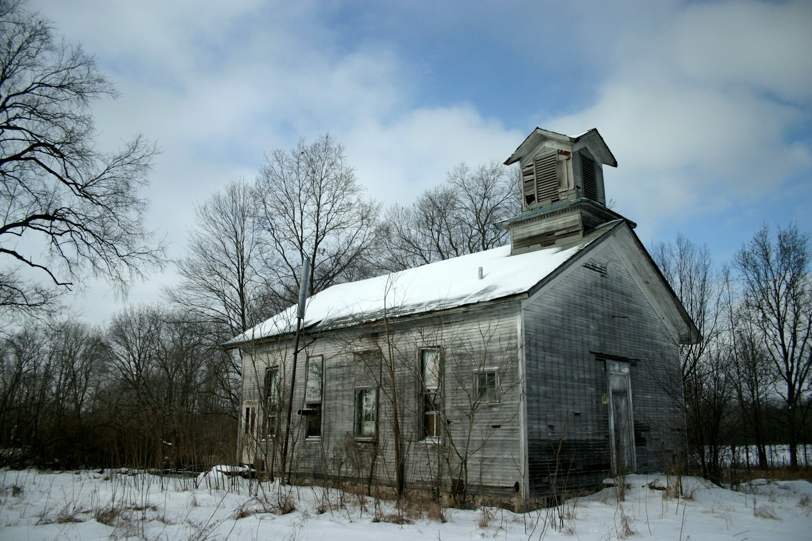 gray wooden house near snowy field surrounded with bare trees under white and blue sky during daytime