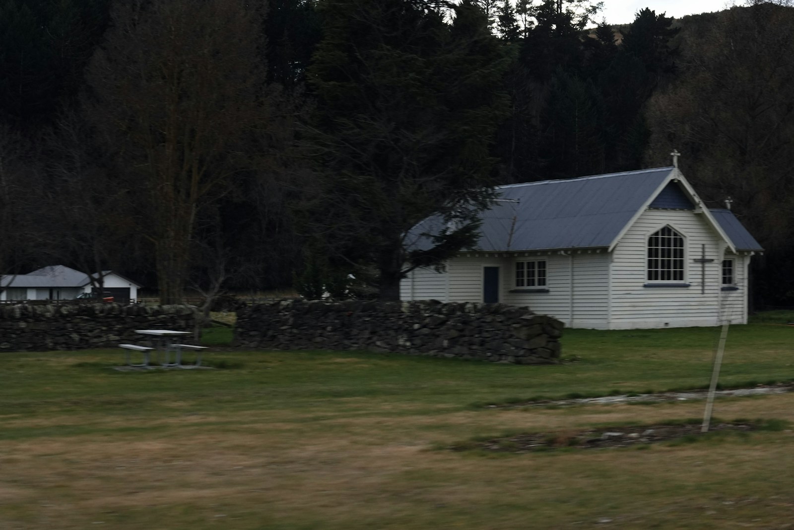 A white wooden church with a grey roof near a stone wall and trees