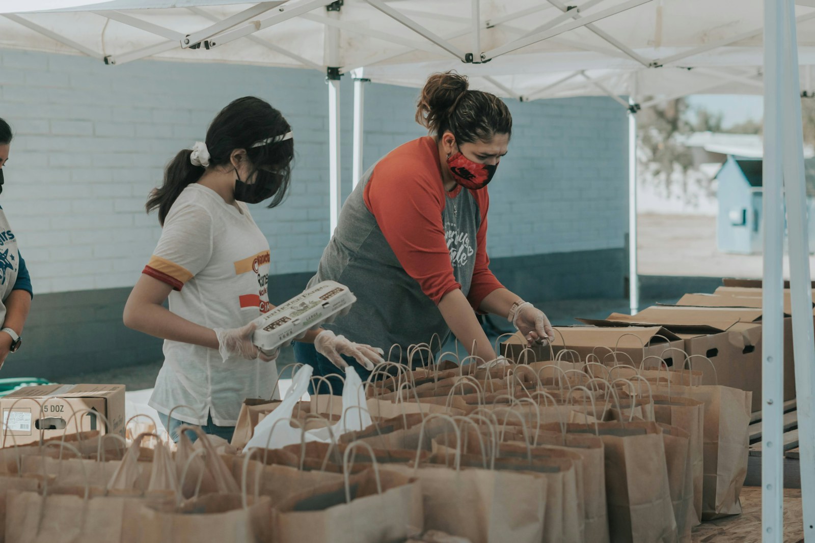 Two women wearing face masks and gloves packing paper bags under a white canopy