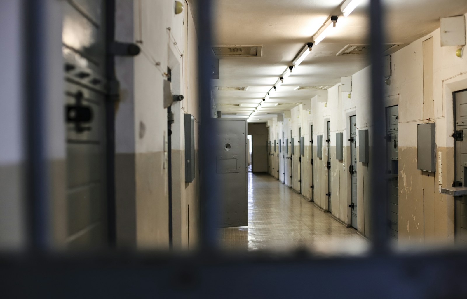 A long prison corridor with rows of metal doors viewed through dark bars